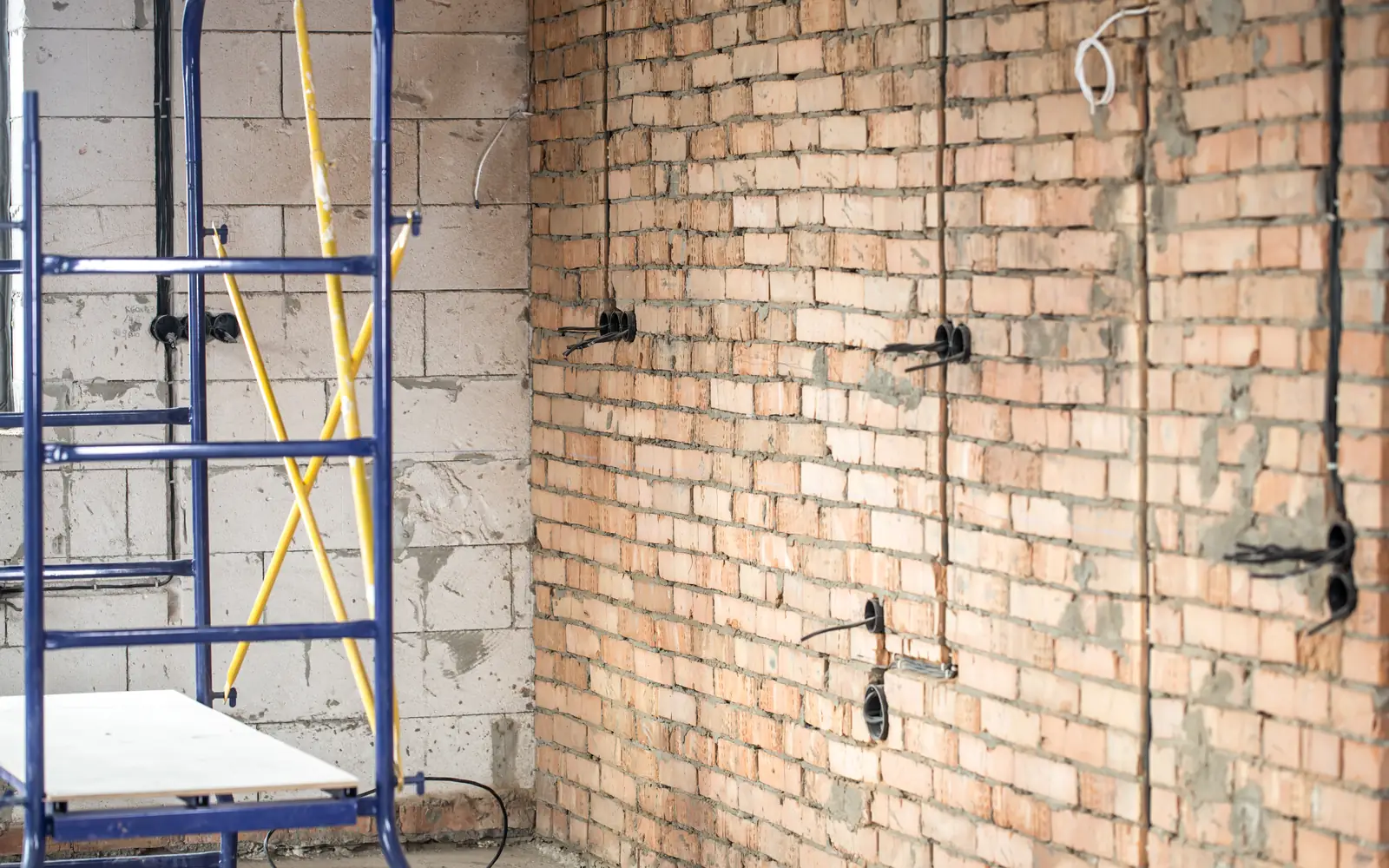 Une pièce en rénovation montre un mur de briques apparent, des gaines électriques et un échafaudage