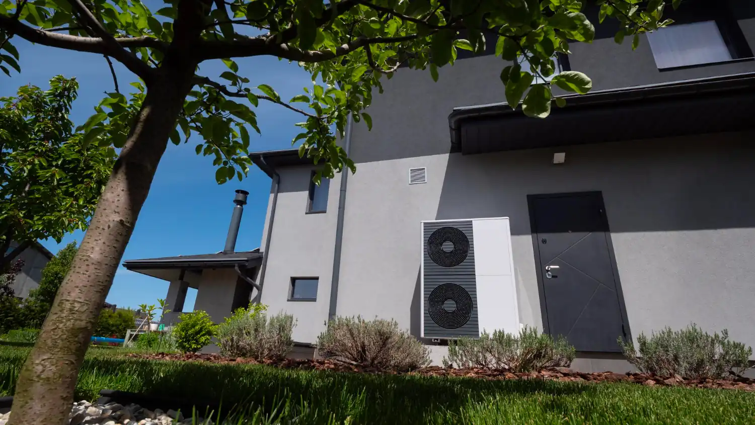 Une unité extérieure de pompe à chaleur est installée près de la façade d’une maison entourée de végétation